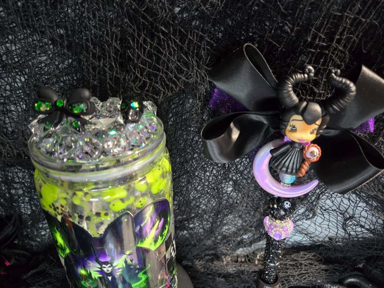 A glass snow globe tumbler filled with slime-green glitter and topped with crushed crystal, a black bow and a skull charm, beside a beaded pen crowned by a horned clay doll on a lavender crescent moon with a black satin bow.