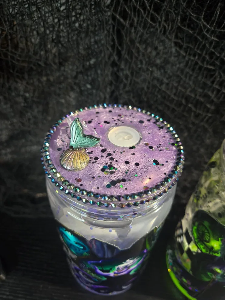 Top-down view of a lavender glitter tumbler lid ringed in iridescent rhinestones, with a gold seashell and teal mermaid-tail charm.