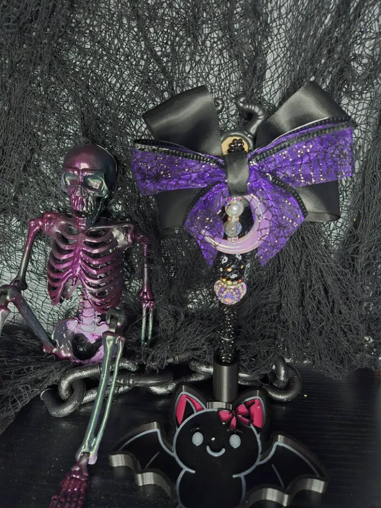 Black beaded pen with a crescent moon charm and a large black and purple spiderweb bow, standing in a bat-shaped holder next to a purple skeleton.