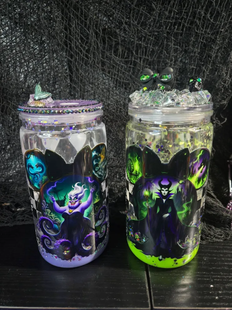 Two snow globe tumblers side by side: a lavender one with a sea-witch print and shell-charm lid, and a slime-green one with a horned sorceress print and crystal lid.