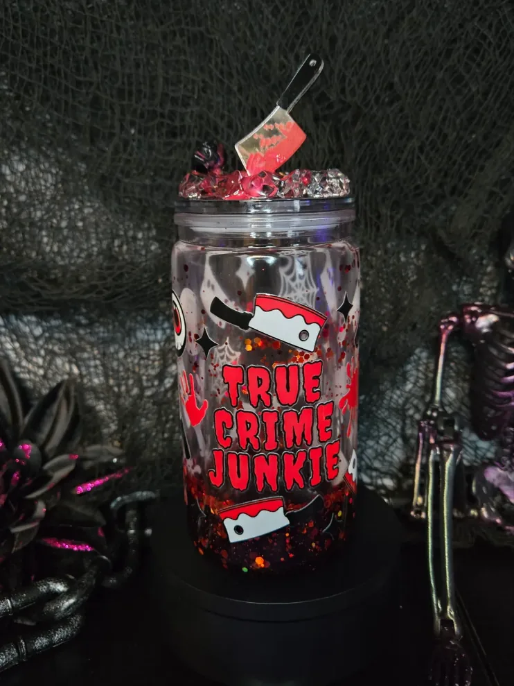 Clear 16 oz snow globe tumbler printed with red cleavers, handprints and the words True Crime Junkie, red glitter settled at the bottom and a bloody cleaver charm in a crystal lid.
